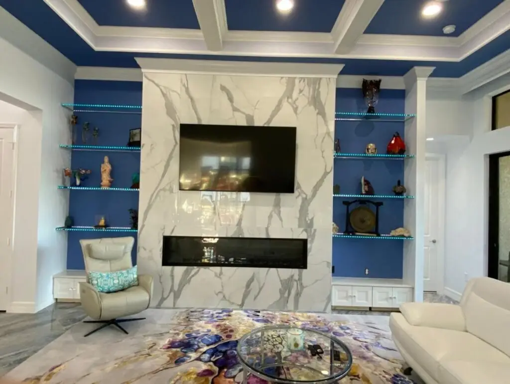 Living room fireplace wall clad in large-format porcelain tile with blue LED display shelving and diagonal 24×48 porcelain flooring at the Lorenzo Residence in Stonecreek Community, Naples, Florida.