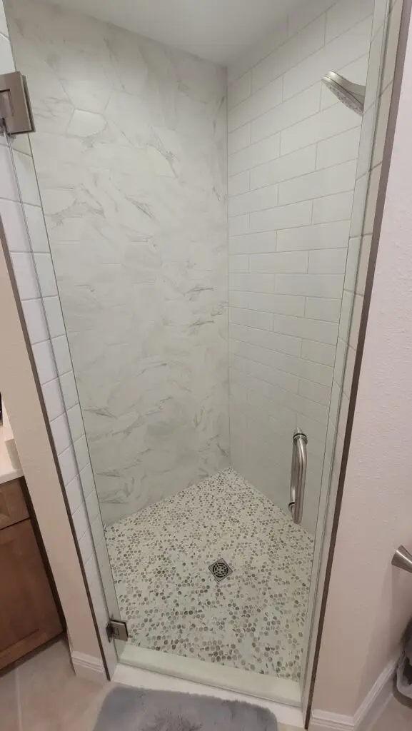 Completed shower from the same Cape Coral bathroom project, with white porcelain wall tile, a frameless glass door, and a round mosaic floor