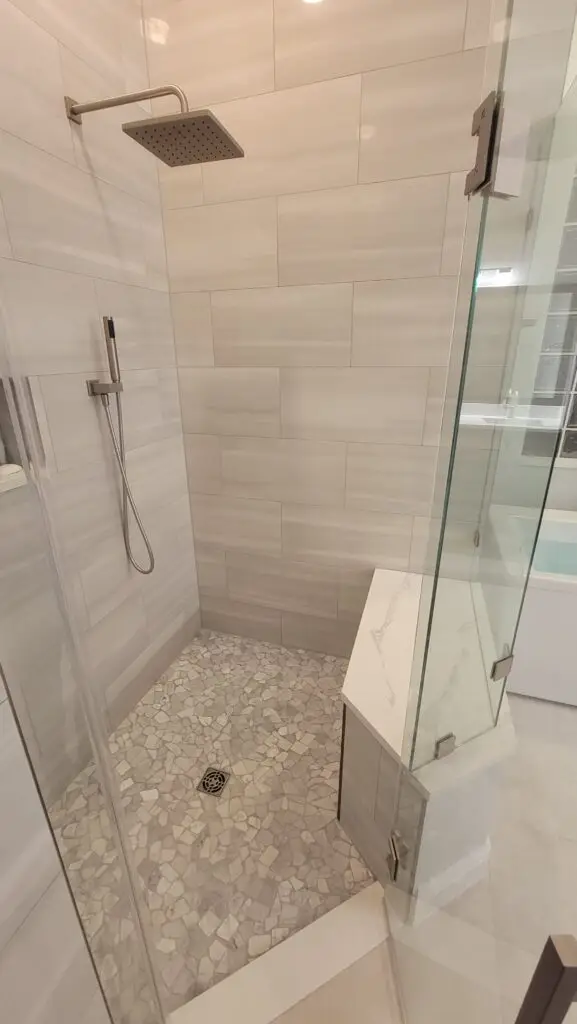Completed porcelain tile shower with frameless glass enclosure, rainfall showerhead, recessed niche, bench, and mosaic floor in Naples.