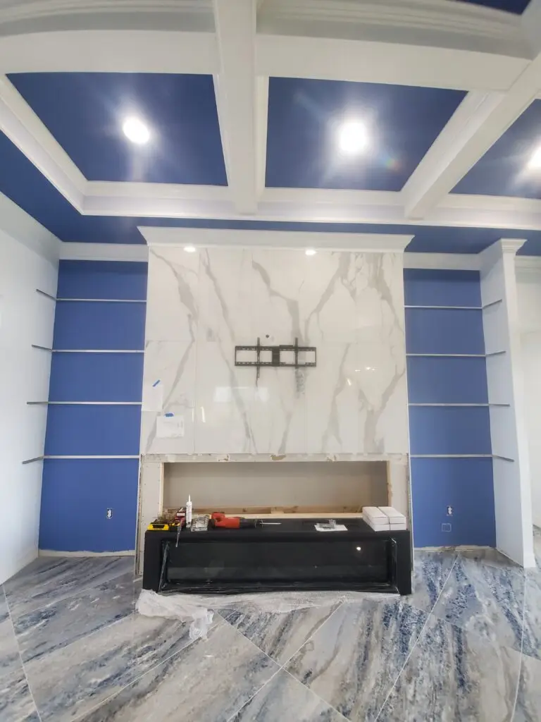 Fireplace feature wall being clad in large-format marble-look porcelain tile between blue display shelving in a Naples home